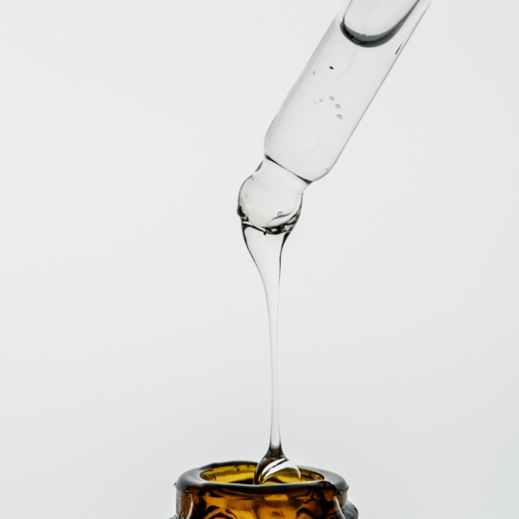 Glass dropper with liquid being dispensed into a amber container on a light gray background