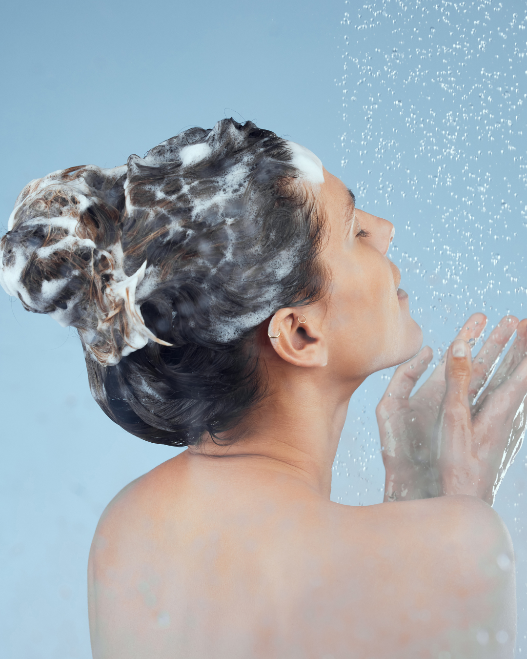 Person with shampoo in hair under a shower against a light blue background